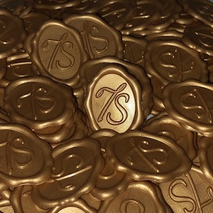 May include: A pile of gold-coloured wax seals. Each seal features an oval design with the letters "TS" in a decorative script. The seals are arranged in a random pattern, creating a textured, layered appearance. The overall effect is elegant and decorative.