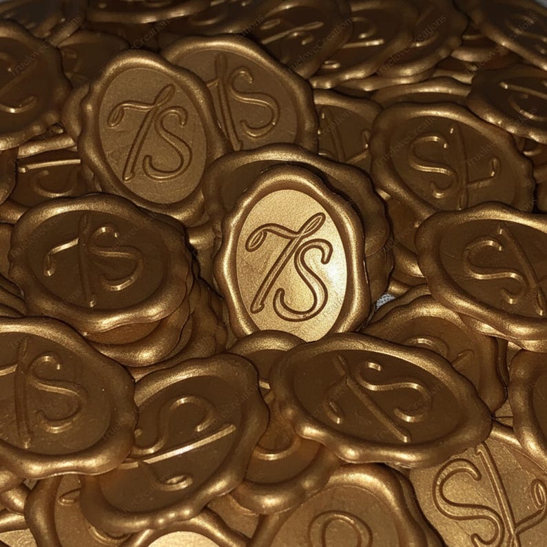 May include: A pile of gold-coloured wax seals. Each seal features an oval design with the letters "TS" in a decorative script. The seals are arranged in a random pattern, creating a textured, layered appearance. The overall effect is elegant and decorative.