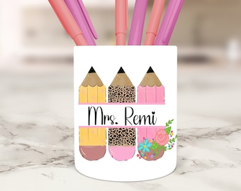 Può includere: Portapenne in ceramica bianca con tre matite, una gialla, una con stampa leopardata e una rosa. Le matite hanno gomme marroni. Il portapenne ha il testo "Mrs. Remi" e fiori rosa.