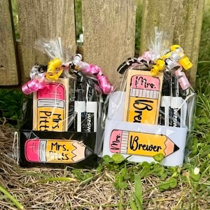 Op de afbeelding: Twee cadeaupakketten met items met een schoolthema. Elk pakket bevat een gum in de vorm van een potlood, twee zwarte stiften en een cadeaulabel in de vorm van een potlood met de naam van een leraar. De pakketten zijn verpakt in cellofaan en vastgebonden met roze en gele linten.