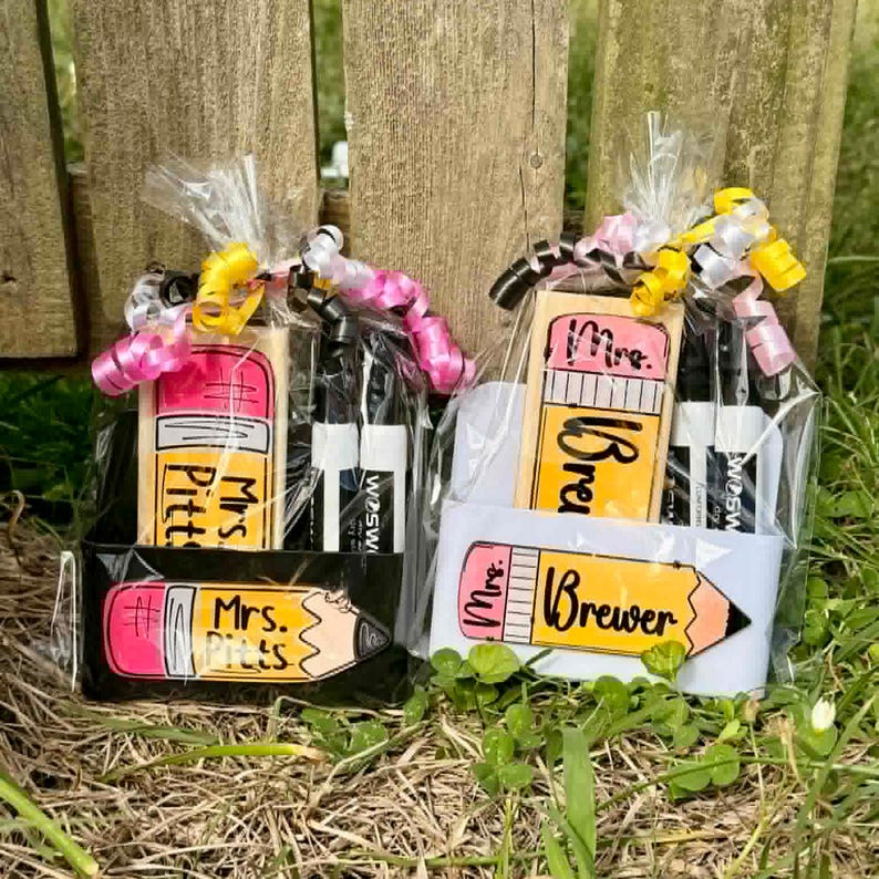 Op de afbeelding: Twee cadeaupakketten met items met een schoolthema. Elk pakket bevat een gum in de vorm van een potlood, twee zwarte stiften en een cadeaulabel in de vorm van een potlood met de naam van een leraar. De pakketten zijn verpakt in cellofaan en vastgebonden met roze en gele linten.