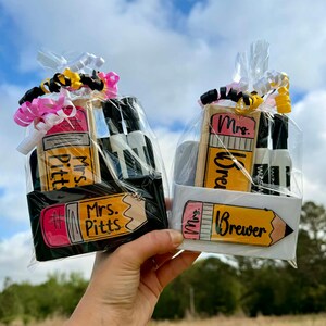 Op de afbeelding: Twee cadeaupakketten, een zwarte en een witte, elk met een potloodvormig item en kleine flesjes. De potloodvormige items hebben de namen "Mrs. Pitts" en "Mrs. Brewer" erop. De pakketten zijn verpakt in doorzichtig cellofaan en vastgebonden met linten.