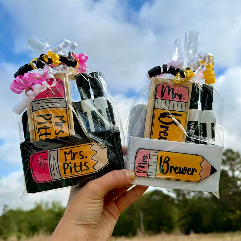 Op de afbeelding: Twee cadeaupakketten, een zwarte en een witte, elk met een potloodvormig item en kleine flesjes. De potloodvormige items hebben de namen "Mrs. Pitts" en "Mrs. Brewer" erop. De pakketten zijn verpakt in doorzichtig cellofaan en vastgebonden met linten.
