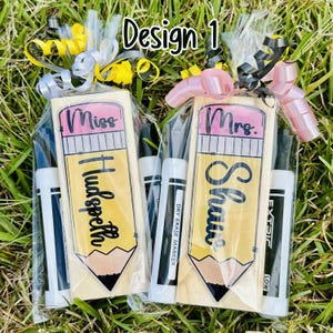 May include: Two teacher gifts with pencil-shaped wooden plaques, personalized with names and titles, packaged with dry-erase markers and ribbons. The text on the plaques reads "Miss Hudspeth" and "Mrs. Shaw". The background is green grass.