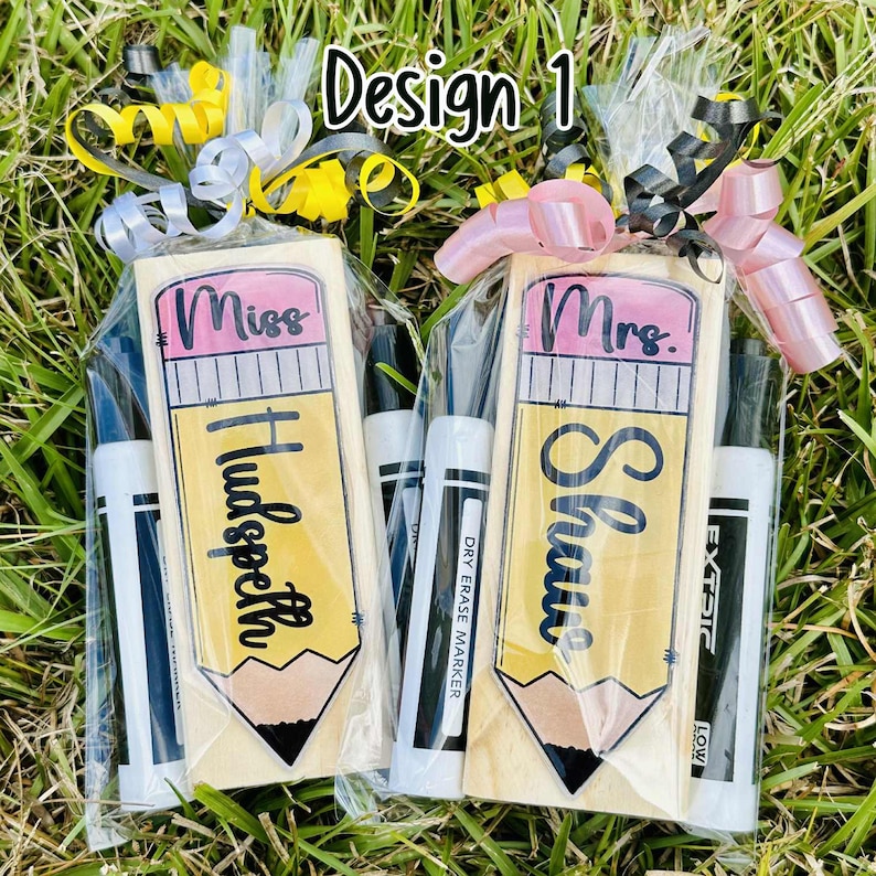 May include: Two teacher gifts with pencil-shaped wooden plaques, personalized with names and titles, packaged with dry-erase markers and ribbons. The text on the plaques reads "Miss Hudspeth" and "Mrs. Shaw". The background is green grass.