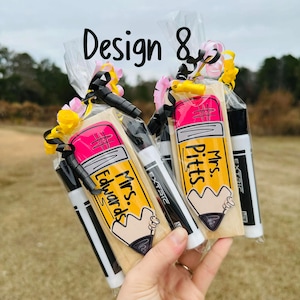 May include: Two gift bags with pencil-shaped cookies, each with a teacher's name, and two markers. The cookies are yellow with pink erasers. The bags are tied with black ribbon and decorated with pink and yellow flowers. The text "Design 8" is at the top.