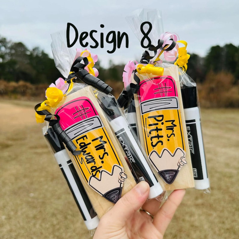 May include: Two gift bags with pencil-shaped cookies, each with a teacher's name, and two markers. The cookies are yellow with pink erasers. The bags are tied with black ribbon and decorated with pink and yellow flowers. The text "Design 8" is at the top.