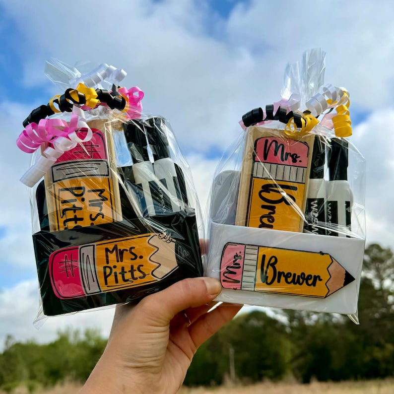 May include: Two gift baskets, one black and one white, each containing a pencil-shaped item and small bottles. The pencil-shaped items have the names "Mrs. Pitts" and "Mrs. Brewer" on them. The baskets are wrapped in clear cellophane and tied with ribbons.