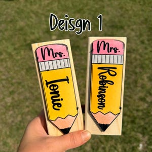 Op de afbeelding: Twee houten naamlabels in de vorm van een potlood. Elk label heeft een geel potloodontwerp met een roze gum en zwarte tekst. Op &eacute;&eacute;n staat "Mrs. Ionie" en op de andere "Mrs. Robinson". De tekst "Design 1" staat erboven.
