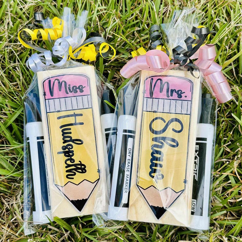 Op de afbeelding: Twee cadeausets voor leraren, elk met een houten potloodvormig bord en droog uitwisbare markers. Op het ene bord staat "Miss Hudspeth" en op het andere "Mrs. Shaw". De cadeaus zijn verpakt in doorzichtig cellofaan en afgewerkt met decoratieve strikken.