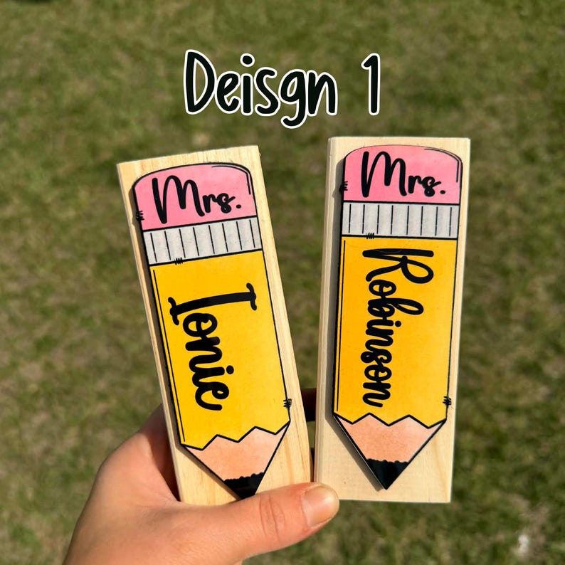 May include: Two wooden pencil-shaped name tags. Each tag features a yellow pencil design with a pink eraser and black text. One reads "Mrs. Ionie" and the other "Mrs. Robinson". The text "Design 1" is above.