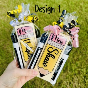 Op de afbeelding: Twee cadeau sets, elk met een potloodvormig item met de tekst "Miss Harper" en "Mrs. Shaw", en een zwart-witte whiteboard marker. De sets zijn verpakt in cellofaan en vastgebonden met linten.