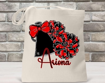 May include: A white canvas tote bag with a black and red cheerleading design. The design includes a black megaphone with a red bow, two red and black pom-poms, and the name "Akiona" in red lettering.