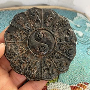 May include: A dark brown, circular pendant featuring a Yin Yang symbol at its centre. The pendant is divided into twelve sections, each with a detailed relief carving of an animal. The pendant is held in a hand.