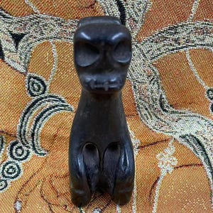 May include: A dark, carved sculpture of a stylized creature with a skull-like face and a squat body. The sculpture is a dark color and has a smooth, polished finish. The piece is likely a decorative item or a small statue.