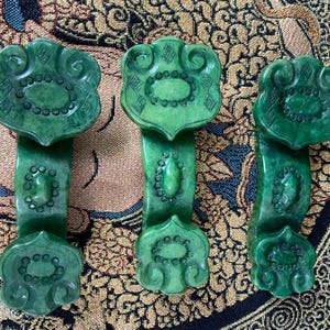 May include: Three carved jade ornaments in a vibrant green hue. Each piece features a stylized floral design at the top and bottom, connected by a central column with a decorative element. The intricate carvings suggest a traditional aesthetic.