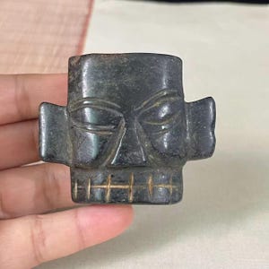 Puede incluir: Un objeto de piedra tallada de color gris oscuro con forma de rostro estilizado, orejas anchas y prominentes, ojos almendrados y una zona bucal rectangular con líneas verticales grabadas rellenas de una sustancia clara.