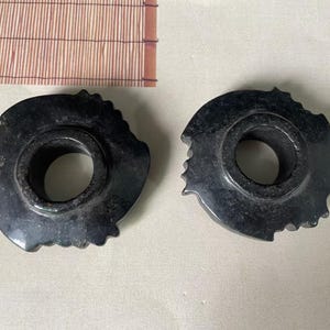 May include: Two circular, dark charcoal-coloured stone objects with a smooth, polished finish. Each piece features a central circular opening and a jagged, uneven outer edge with multiple small, protruding points along the perimeter.