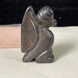 May include: A dark brown, carved stone figurine depicting a stylized creature with a prominent wing, a rounded head, and a seated posture. The surface of the stone has a textured, matte appearance with subtle variations in color and natural markings.