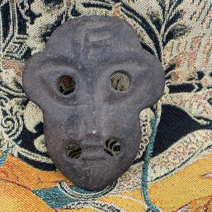May include: A dark grey mask-shaped object featuring two eye holes and two mouth holes. The mask has a slightly rough texture, set against a patterned fabric background with yellow, black, and teal colours.
