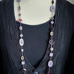 May include: A long purple and brown beaded necklace with various shapes and sizes of beads. The necklace is on a black shirt.