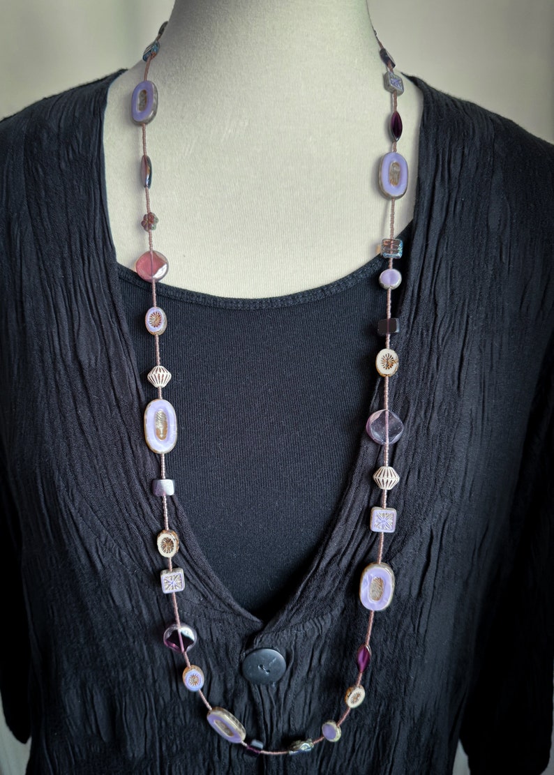 May include: A long purple and brown beaded necklace with various shapes and sizes of beads. The necklace is on a black shirt.