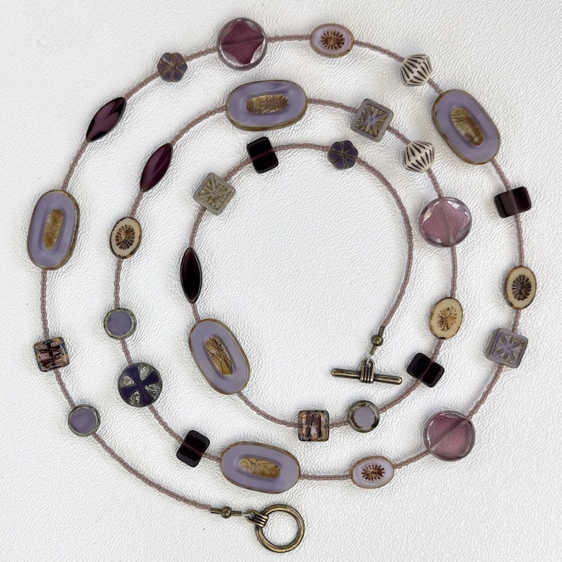 May include: A long, layered necklace with a variety of purple and brown glass beads. The beads are in different shapes, including squares, ovals, and circles. The necklace has a silver clasp.