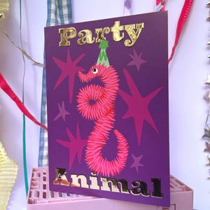 Op de afbeelding: Een paarse feestuitnodigingskaart met gouden folieletters met de tekst "Party Animal". De kaart toont een roze worm met een feesthoed en roze sterren. Decoratieve linten en sterrenslingers op de achtergrond.