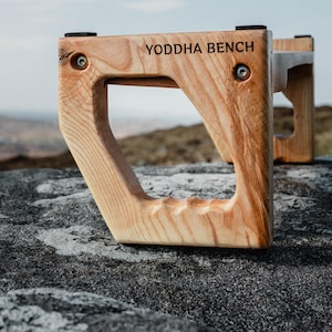 May include: A wooden bench with the words "YODDHA BENCH" engraved on the top. The bench is made of light brown wood and has a natural finish. The bench is sitting on a gray rock.