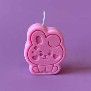 May include: A pink wax candle shaped like a cartoon rabbit with a white wick at the top. The rabbit design features embossed details including two heart shapes on the cheeks, a small nose, a mouth, and a four-pointed star icon on the left side.