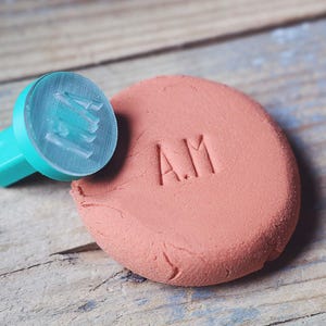 May include: A teal stamp with the initials "AM" imprinted on a round of pink clay. The stamp is next to the clay, which is resting on a wooden surface. The letters are clearly visible.