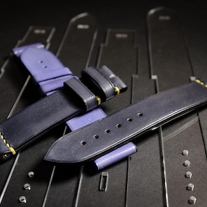 Op de afbeelding: Twee marineblauwe leren horlogebandjes met gele stiksels en zilveren hardware. De bandjes worden getoond op een donkere ondergrond met horlogemakerssjablonen. Eén bandje is gesloten, de andere is open.