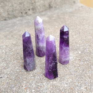 May include: Four amethyst crystal points, each with a different shade of purple, are arranged on a gray concrete surface. The points are all roughly the same size and shape, and they have a natural, unpolished finish.