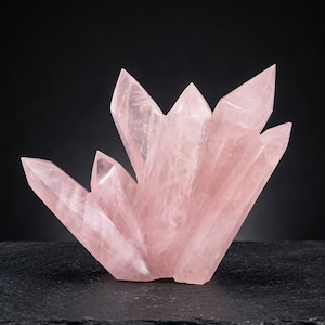 Pode incluir: Um aglomerado de cristais de quartzo rosa pálido com múltiplas terminações afiadas e pontiagudas. A formação mineral translúcida exibe fraturas internas naturais e tons variados de rosa suave em toda a sua estrutura cristalina irregular.