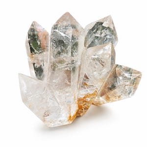 May include: A cluster of clear quartz crystals with sharp, pointed terminations. The crystals have internal inclusions and a slightly translucent appearance. The base of the cluster has a light brown hue, and the crystals are set against a white background.