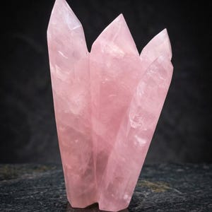 Puede incluir: Un grupo de cristales de cuarzo rosa translúcido con múltiples terminaciones puntiagudas. El espécimen mineral muestra una suave coloración rosa pálido con fracturas internas naturales y niveles variables de claridad en toda la estructura vertical.