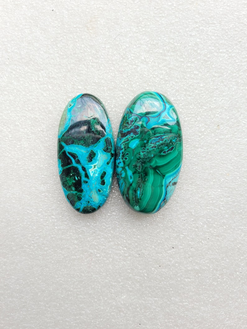Op de afbeelding: Twee ovale cabochons van groene en blauwe chrysocolla. De stenen hebben een draaiende patroon van groen, blauw en zwart.