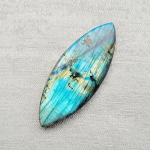 May include: A blue and green labradorite gemstone cabochon with iridescent shimmer. The oval-shaped stone has a smooth, polished surface.