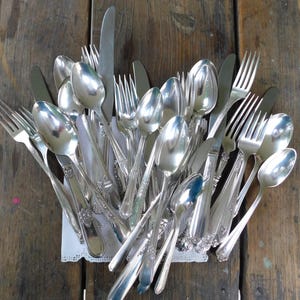 May include: A collection of vintage silverware, including forks, spoons, and a knife, arranged in a fan shape on a white lace doily. The silverware is made of polished silver and has a classic design.
