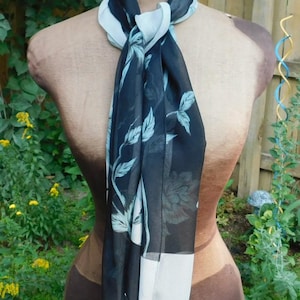 May include: A black and white floral patterned scarf with a white trim. The scarf is tied around a mannequin's neck.