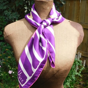 May include: A purple and white striped silk scarf with a pink border. The scarf has the word "Una" printed on it.