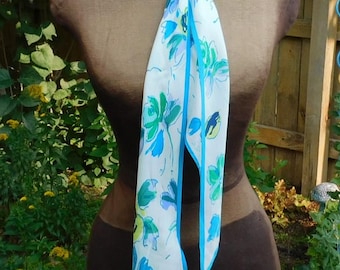 May include: A white silk scarf with a blue and green floral pattern. The scarf is tied around a mannequin's neck and is hanging down in front of the mannequin.