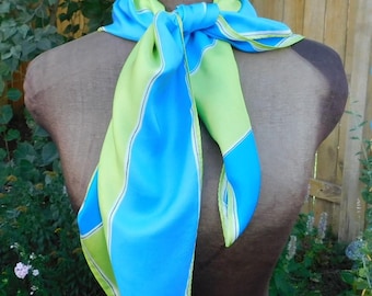 May include: A silk scarf in blue and green hues is displayed on a mannequin. The scarf has a colour block design with a light green border and a bright blue centre. The scarf is knotted at the neck, highlighting its flowing fabric.