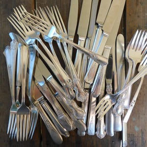 May include: A collection of vintage silverware, including forks and knives, in a silver finish. The silverware is arranged in a pile, with the handles pointing in different directions.