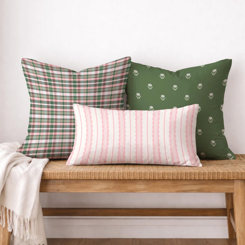 May include: Three decorative throw pillows on a wooden bench. One pillow has a green and pink plaid pattern, another is green with a white floral design, and the third is pink and white striped.