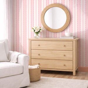 May include: A light wood dresser with three drawers, a round mirror with a woven frame, and a white armchair. The room has pink and white striped wallpaper and a woven basket.