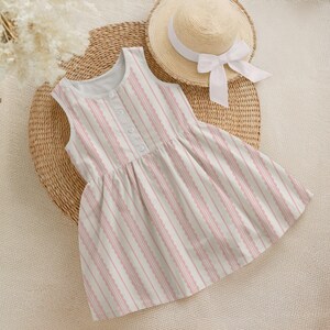 May include: A sleeveless dress with a pink and white striped pattern and a row of buttons down the front. A straw hat with a white ribbon sits nearby. The dress is on a woven mat.