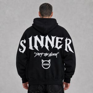 Może przedstawiać: Czarna bluza z kapturem z napisem SINNER wykonanym dużymi, białymi literami w stylu gotyckim na plecach. Poniżej tekstu znajduje się napis Defy the Norm białą czcionką oraz biała okrągła ikona z rogami diabła i trzema znakami X w środku.