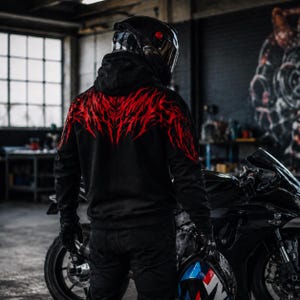 May include: A black hooded sweatshirt featuring a vibrant red tribal-style graphic pattern across the upper back and shoulders. The garment has long sleeves, a hood, and a solid black lower section, worn with black trousers and black gloves.