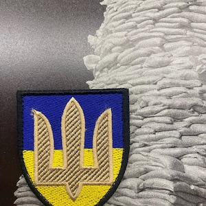 Puede incluir: Parche bordado con el escudo de armas ucraniano, un tridente, en hilo dorado. El parche tiene una sección superior azul y una sección inferior amarilla, con un borde negro. El fondo es una imagen en escala de grises de sacos de arena.